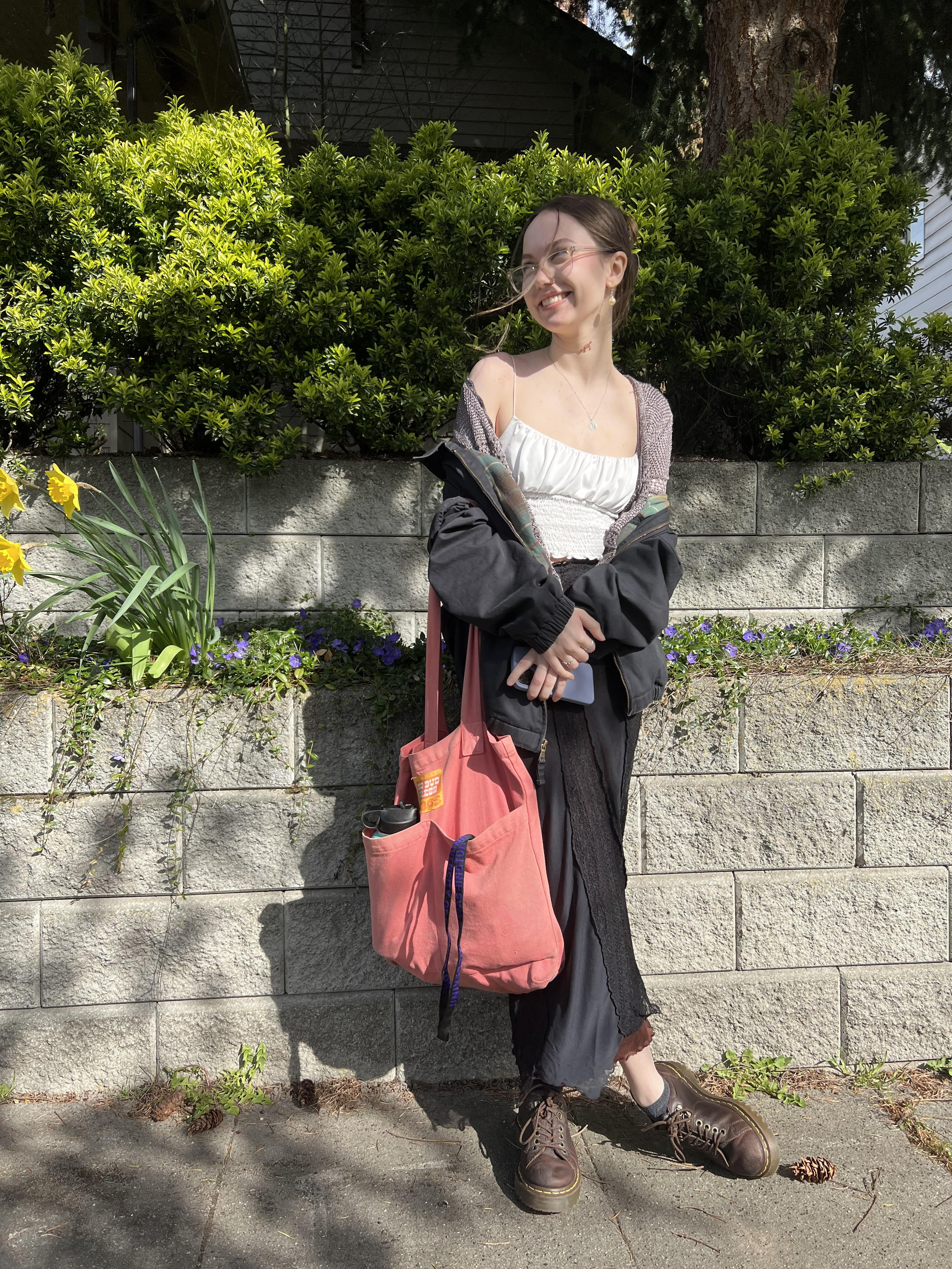 photo of white girl with brown hair, wearing a jacket, a white tank top, and a skirt, carrying a pink bag.